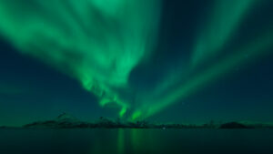 Powerful aurora borealis lighting up the night sky above Korsnes, Norway, reflected over calm water in February